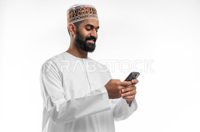إستخدام الموبايل لإنجاز المهام,  بورتريه مقرب لرجل عربي خليجي عماني مبتسم يرتدي الدشداشة و الكمه يحمل الهاتف المحمول بيده بإيماءات السعادة و السرور، تطور التكنولوجيا والأجهزة التقنية الحديثة, خلفية بيضاء