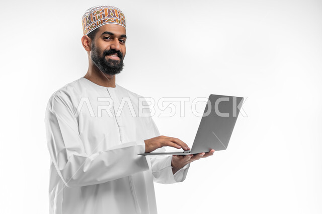 دمج التقنيات الحديثة بالحياة اليومية, مفهوم العمل و انجاز المهام عن بعد, بورتريه مقرب لرجل عربي خليجي عماني مبتسم يرتدي الدشداشة و الكمه يحمل بيده جهاز الحاسوب المحمول و ينظر الى الكاميرا بتعابير السرور, خلفية بيضاء