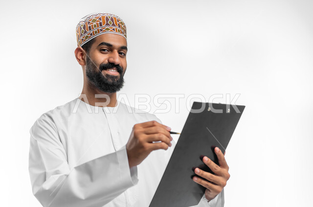 تعابير التفكير لاتخاذ القرارات, تدوين و تسجيل الملاحظات, بورتريه مقرب لرجل عربي عماني خليجي يرتدي الدشداشة و الكمه يمسك بيديه مفكرة وقلم يقوم بكتابة المعلومات, النظر الى الكاميرا بإيماءات التفاؤل و السرور، خلفيه بيضاء