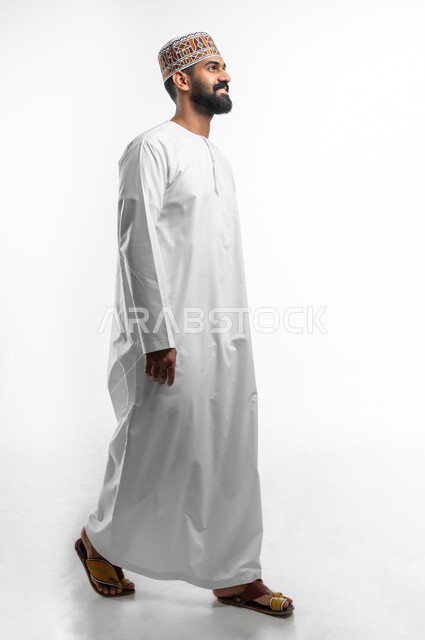 The concept of elegance and attention to external appearance, a portrait from the side of a young Gulf Arab man wearing a dishdasha and a cuff with gestures of happiness and pleasure, expressions of walking and walking to go somewhere, full-length body image, length, white background