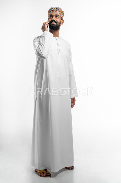 استخدام التقنيات الحديثة المتطورة, ايماءات السعادة و السرور، بورتريه لشاب عربي خليجي عماني يرتدي الدشداشه و الكمه يقوم بإجراء مكالمة هاتفية عبر الهاتف المحمول، التواصل مع الاهل و الاصدقاء, صورة الجسم بالطول الكامل, خلفية بيضاء