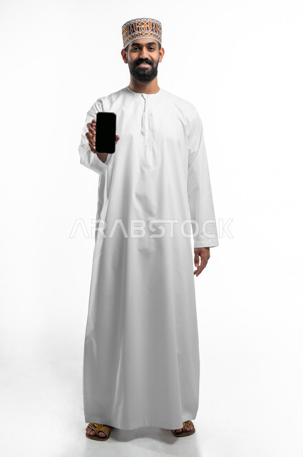 استخدام التكنولوجيا و التقنيات الحديثة المتطورة، بورتريه لشاب عربي خليجي عماني مبتسم يرتدي الدشداشة و الكمه يقوم بعرض شاشة سوداء فارغة عبر الهاتف المحمول،النظر الى الكاميرا بتعابير السرور، صورة الجسم بالطول الكامل, خلفية بيضاء