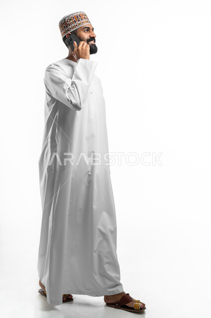 Gestures of happiness and pleasure, a portrait of a young Arab Gulf Omani man wearing a dishdasha and cuff making a phone call via a mobile phone, using advanced modern technologies, communicating with family and friends, full-length body photo, white background.