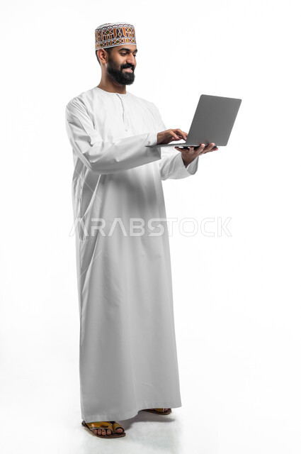 مفهوم العمل و انجاز المهام عن بعد, دمج التقنيات الحديثة بالحياة اليومية, بورتريه لرجل عربي خليجي عماني مبتسم يرتدي الدشداشة و الكمه يحمل بيده جهاز الحاسوب المحمول، الجسم بالطول الكامل, خلفية بيضاء