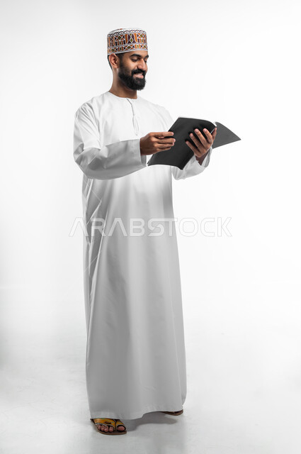 التزود بالعلم والمعرفة، قضاء وقت الفراغ بالتثقيف والتعلم، بورتريه لشاب عربي خليجي عماني يرتدي الدشداشة و الكمه يمسك كتابا بيده يقوم بالقراءه بايماءات الفرح و السرور، الجسم بالطول الكامل, خلفية بيضاء
