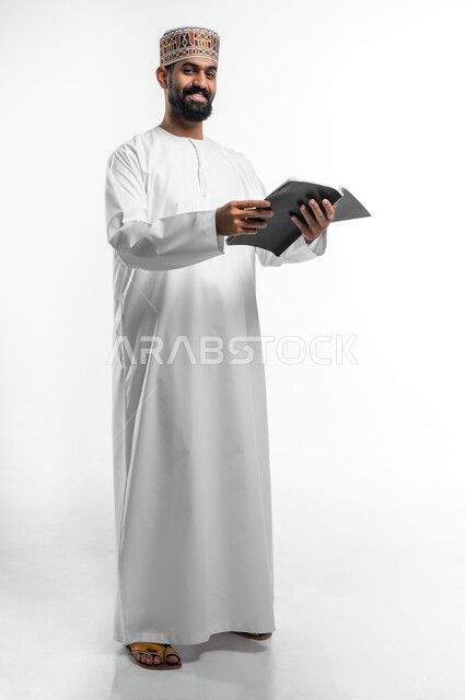 النظر الى الكاميرا بايماءات الفرح و السرور, ممارسة هواية القراءة و التزود بالعلم، قضاء وقت الفراغ بالتثقيف والتعلم، بورتريه لشاب عربي خليجي عماني يرتدي الدشداشة و الكمه يمسك بيده كتابا علميا، الجسم بالطول الكامل, خلفية بيضاء
