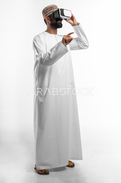 المتعة و التسلية بالمشاهدة عبر نظارة الواقع الافتراضي, بورتريه لشاب عربي خليجي عماني يرتدي نظارات ثلاثية الأبعاد, الاندماج ومحاكاة العالم الخيالي, محاولة رفع اصبع السبابة بتعابير محاولة النقر على شيء ما, صورة الجسم بالطول الكامل, خلفية بيضاء