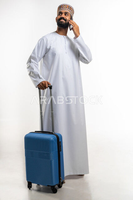 التواصل مع الاصدقاء و الاقارب عبر الهاتف المحمول بتعابير السرور, قضاء أوقات ممتعة في الإجازة, بورتريه شاب عربي خليجي عماني يمسك حقيبة السفر يرتدي الدشداشة و الكمه يقوم باجراء مكالمة هاتفية عبر الجوال, الجسم بالطول الكامل, خلفية بيضاء