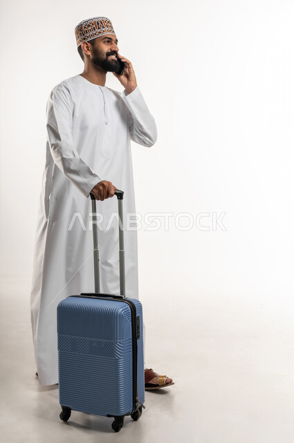 قضاء أوقات ممتعة في الإجازة, بورتريه شاب عربي خليجي عماني يمسك حقيبة السفر يرتدي الدشداشة و الكمه يقوم باجراء مكالمة هاتفية عبر الجوال, التواصل مع الاصدقاء و الاقارب عبر الهاتف المحمول بتعابير السرور, الجسم بالطول الكامل, خلفية بيضاء