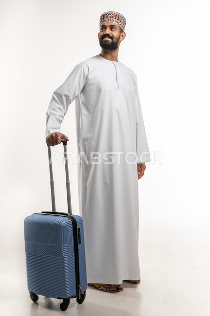 الاستعداد لقضاء رحلة ممتعة في الاجازةت الصيفية ، مفهوم السياحة و السفر, بورتريه لشاب عربي خليجي عماني يرتدي الدشداشة و الكمه يمسك بيده حقيبة الأمتعة و ينظر الى شيئ ما بتعابير السرور, الجسم بالطول الكامل، خلفية بيضاء