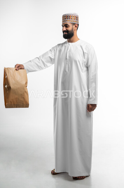 خدمة توصيل الطلبات و البضائع الى الزبائن, بورتريه لشاب عربي خليجي عماني يرتدي الدشداشة و الكمه يقوم بتسليم طرد بريدي الى الزبون,خدمات الديليفري السريعة و الآمنة ذات الجودة العالية, الجسم بالطول الكامل, خلفية بيضاء