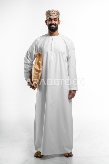 Delivery and receipt of electronic orders, a portrait of a smiling Arab Gulf Omani man wearing a dishdasha and a cuff, holding a postal parcel under his arms, looking at the camera with gestures of happiness and pleasure, full-length body portrait, white background.
