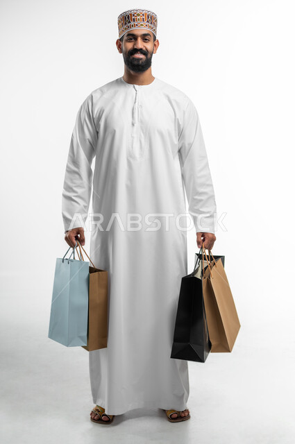 الاستفادة من العروض و التخفيضات الموسمية في الاسواق,  بورتريه لشاب عربي خليجي عماني مبتسم يرتدي الدشداشة والكمه يحمل بيده اكياس التسوق الملونة ينظر الى الكاميرا بايماءات السرور, متعة في التسويق و اقتناء المستلزمات و الهدايا, الجسم بالطول الكامل, خلفيه بيضاء