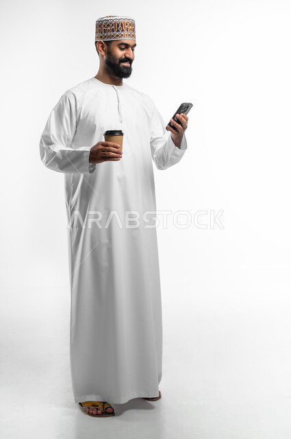 استخدام التقنيات الحديثة المتطورة, ضمان توصيل المشروبات الساخنة, بورتريه لشاب عربي خليجي عماني يرتدي الدشداشة والكمه يحمل بيده كوب من القهوة و الهاتف، طلبات الاونلاين السريعة و الآمنة, الجسم بالطول الكامل, خلفيه بيضاء