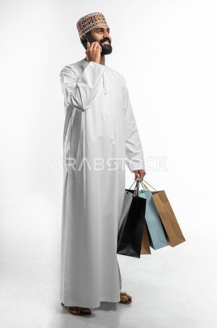 التواصل مع العميل للتأكد من الموقع, دمج التقنيات الحديثة بالحياة العملية, بورتريه لشاب عربي خليجي عماني مبتسم يرتدي الدشداشة والكمه يحمل بيده الأكياس الورقية و يقوم باجراء مكالمة عبر الهاتف المحمول ، صورة الجسم بالطول الكامل, خلفيه بيضاء