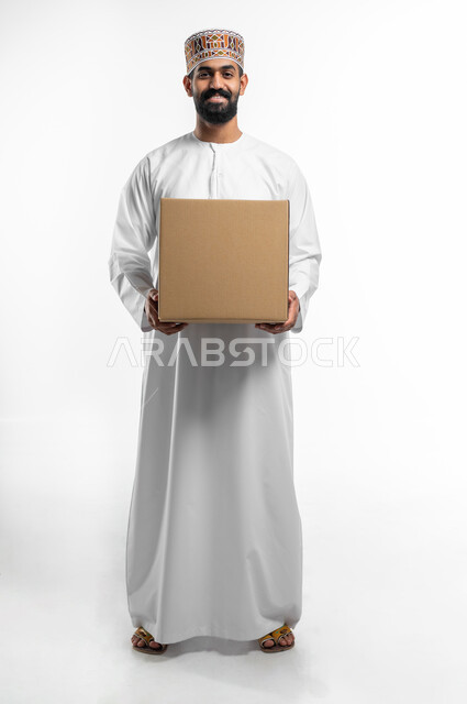Looking at the camera with confidence and happiness for the order to arrive quickly, delivering orders on time and properly, a close-up portrait of an Arab Gulf Omani young man wearing a dishdasha and a sleeve, holding a closed cardboard box in his hand, full-length body photo, white background.