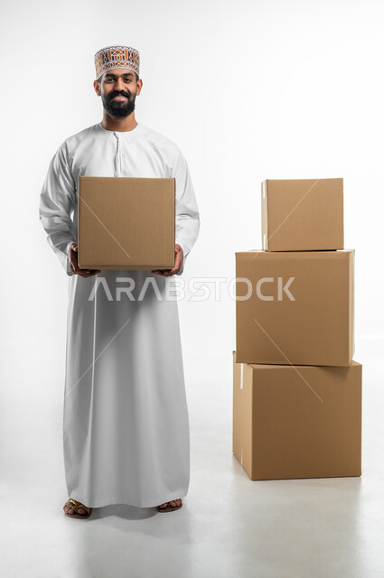 إستلام المشتريات و تسليم البضائع للزبائن, توصيل الطلبيات بالوقت المحدد و بشكل سليم, بورتريه لرجل عربي خليجي عماني يرتدي الكمه و الدشداشة يحمل بيده صندوق كرتوني مغلق, صورة الجسم بالطول الكامل, خلفيه بيضاء