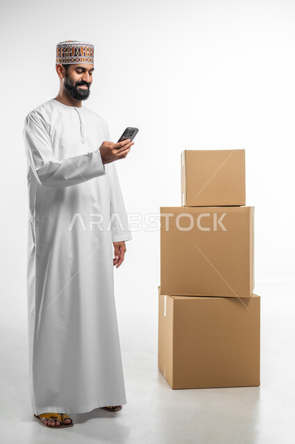 الحجز و الطلب عبر الموبايل, مفهوم التسوق الالكتروني, بورتريه لرجل عربي خليجي عماني يرتدي الدشداشة و الكمه يحمل بيده الهاتف المحمول, التواصل و الدردشة مع العملاء, صورة الجسم بالطول الكامل, خلفيه بيضاء