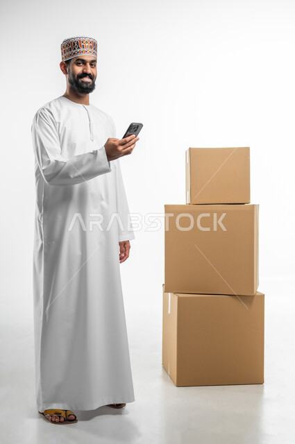 تاكيد الحجوزات عبرالاجهزة التقنية الحديثة, مفهوم التسوق الالكتروني, بورتريه لرجل عربي خليجي عماني يرتدي الدشداشة و الكمه يحمل بيده الهاتف المحمول و ينظر الى الكاميرا بتعابير السرور, التواصل و الدردشة مع العملاء, صورة الجسم بالطول الكامل, خلفيه بيضاء