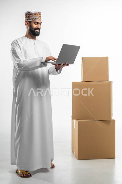 التواصل و الدردشة مع العملاء, مفهوم التسوق الالكتروني, بورتريه لرجل عربي خليجي عماني يرتدي الدشداشة و الكمه يحمل بيده جهاز الحاسوب المحمول و ينظر الى الكاميرا بتعابير السرور, تاكيد الحجوزات عبرالاجهزة التقنية الحديثة, صورة الجسم بالطول الكامل, خلفيه بيضاء
