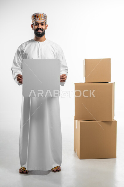 الوقوف بجانب صناديق طلبات مشتريات الاونلاين, بورتريه لشاب عربي خليجي عماني يرتدي الدشداشه و الكمه يحمل بيده لوحة بيضاء مربعة فارغة و ينظر الى الكاميرا بتعابير السرور, موك اب مربع ابيض, اعلانات و عروض تسويقية، الجسم بالطول الكامل, خلفيه بيضاء