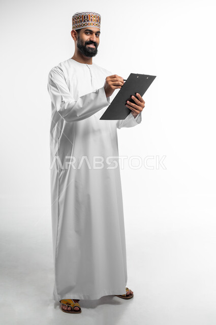 تعابير التفكير لاتخاذ القرارات, تدوين و تسجيل الملاحظات, بورتريه لرجل عربي عماني خليجي يرتدي الدشداشة و الكمه يمسك بيديه مفكرة وقلم يقوم بكتابة المعلومات, النظر الى الكاميرا بإيماءات التفاؤل و السرور، خلفيه بيضاء