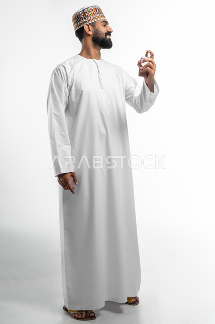 الاستعداد للذهاب والخروج من المنزل, التسويق للعطور الخليجية العمانية, استخدام الزيوت العطرية الطبيعية, بورتريه من الجانب لرجل عربي خليجي عماني مبتسم يرتدي الدشداشه و الكمه يقوم برش العطر بتعابير السرور, صورة الجسم بالطول الكامل, خلفية بيضاء