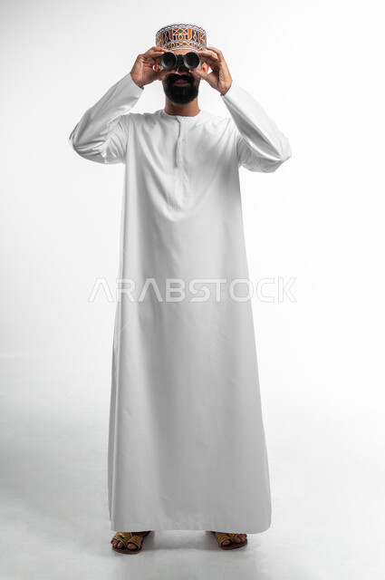 حب الاستكشاف و المغامرة, رؤية الاشياء البعيدة و تقريبها, بورتريه لشاب عربي خليجي عماني يرتدي الدشداشة و الكمه مبتسم يحمل بيده منظار ثنائي العينين و يينظر الى شيئ ما, ايماءات الاستمتاع و السرور, صورة الجسم بالطول الكامل, خلفية بيضاء