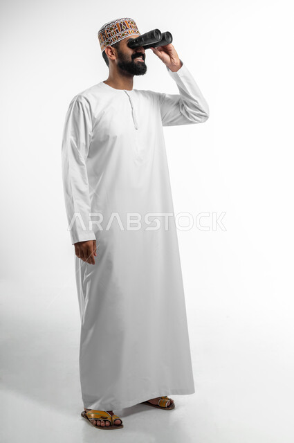 رؤية الاشياء البعيدة عبر التلسكوب, حب الاستكشاف و المغامرة, بورتريه من الجانب لشاب عربي خليجي عماني مبتسم يرتدي الدشداشة و الكمه يحمل بيده منظار ثنائي العينين و يينظر الى شيئ ما, ايماءات الاستمتاع و السرور, صورة الجسم بالطول الكامل, خلفية بيضاء
