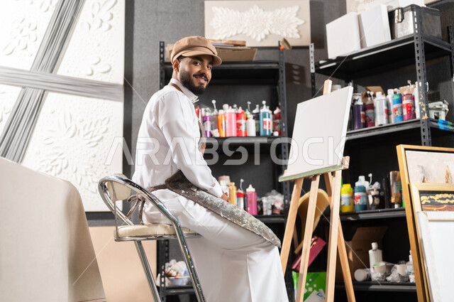 الشغف في ممارسة هواية الرسم و الفنون اليدوية , أنشطة ترفيهية ممتعة , ورشة عمل فنية , رسام عربي خليجي سعودي يرتدي الثوب التقليدي و مريلة الرسم يجلس على الكرسي أمام اللوحة , النظر إلى الكاميرا بإيماءات السعادة و السرور