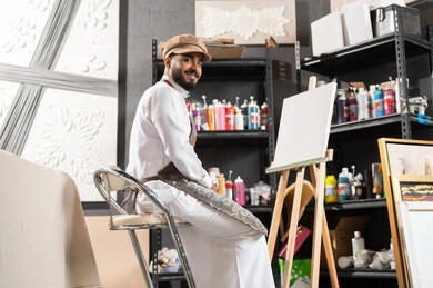 Passion for practicing the hobby of drawing and hand arts, fun recreational activities, an art workshop, a Saudi Arabian Gulf painter wearing a traditional dress and a drawing apron, sitting on the chair in front of the painting, looking at the camera with gestures of happiness and pleasure.