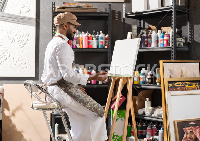 الموهبة و الإبداع في الفنون اليدوية , الشغف في ممارسة الهوايات المفضلة , استخدام الريشة للرسم على اللوحة , ورشة عمل فنية , رسام عربي خليجي سعودي يرتدي الثوب التقليدي و مريلة الرسم يجلس على الكرسي يفكر في رسم شيء ما