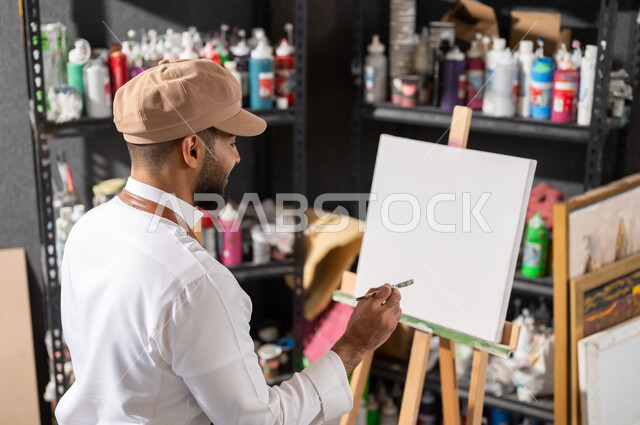ورشة عمل فنية , استخدام الريشة للرسم على اللوحة , الشغف في ممارسة الهوايات المفضلة و الفنون اليدوية , رسام عربي خليجي سعودي يرتدي الثوب التقليدي و مريلة الرسم يجلس على الكرسي يفكر في رسم شيء ما