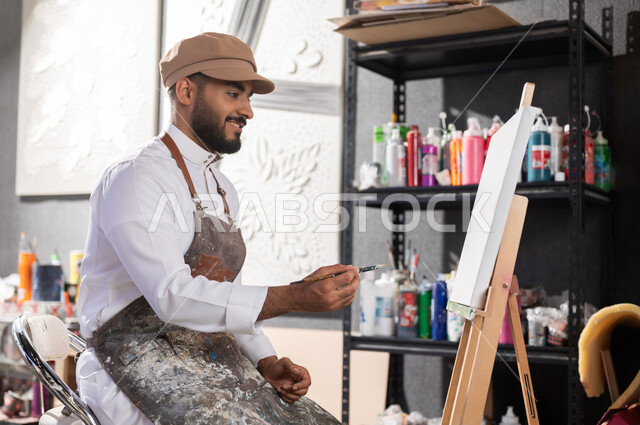 Using a brush to draw on a canvas, an art workshop, a passion for practicing favorite hobbies and handmade arts, a Saudi Gulf Arab painter wearing a traditional dress and a drawing apron sitting on a chair thinking about drawing something