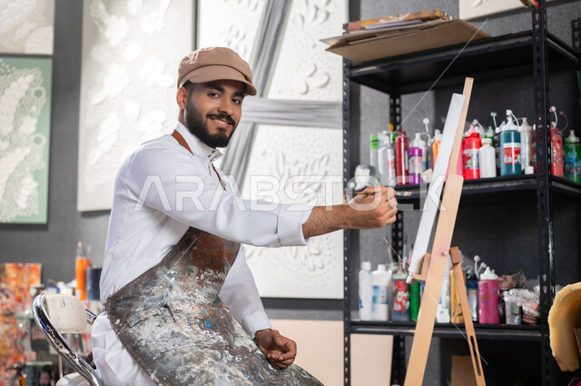 استخدام أدوات و معدات التلوين , إتقان هواية الفنون اليدوية , ورشة عمل فنية , رسام عربي خليجي سعودي يرتدي الثوب التقليدي و مريلة الرسم يجلس على الكرسي أمام اللوحة , النظر إلى الكاميرا بإيماءات الفرح و السرور