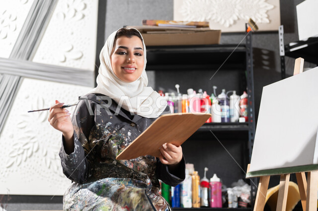 استخدام أدوات و معدات التلوين , ورشة عمل فنية , الشغف في ممارسة الهوايات المفضلة و الفنون اليدوية , رسامة عربية خليجية سعودية محجبة ترتدي مريلة الرسم تجلس على الكرسي , النظر إلى الكاميرا بإيماءات الفرح و السرور