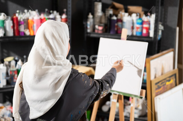 إتقان هواية الفن التشكيلي , الشغف في ممارسة الهوايات المفضلة و الفنون اليدوية , صورة من الخلف لرسامة عربية خليجية سعودية محجبة ترتدي مريلة الرسم تمسك الريشة بيدها , مفهوم الموهبة و الإبداع , ورشة عمل فنية