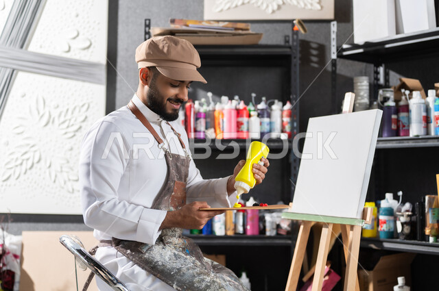 مفهوم الموهبة و الإبداع , ورشة عمل فنية , رسام عربي خليجي سعودي يرتدي الثوب التقليدي و مريلة الرسم يقوم باستعمال اللون الأصفر , الشغف في ممارسة الهوايات المفضلة , استخدام أدوات و معدات الرسم و التلوين