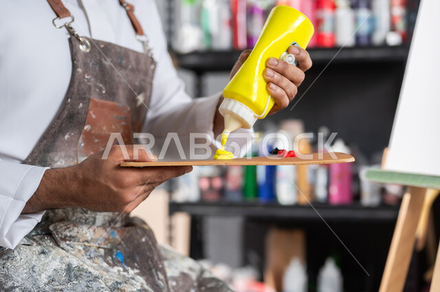 الشغف في ممارسة الهوايات المفضلة , استخدام أدوات و معدات الرسم و التلوين , صورة مقربة لرسام عربي خليجي سعودي يرتدي الثوب التقليدي و مريلة الرسم يقوم باستعمال اللون الأصفر , مفهوم الموهبة و الإبداع , ورشة عمل فنية