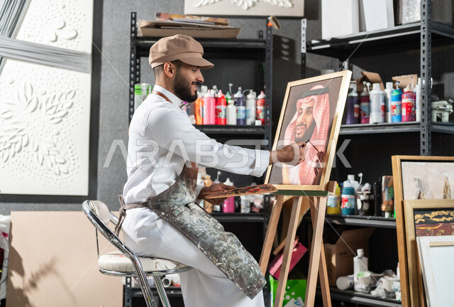الموهبة و الإبداع في الفنون اليدوية , مفهوم الحب و الانتماء للوطن العربي السعودي , ورشة عمل فنية , رسام عربي خليجي سعودي يرتدي الثوب التقليدي و مريلة الرسم يقدم فنون الرسم و التلوين لصورة ولي العهد محمد بن سلمان