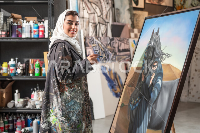 النظر إلى الكاميرا بإيماءات الفرح و السرور , ممارسة هواية الفنون اليدوية , رسامة عربية خليجية سعودية محجبة ترتدي مريلة الرسم تقف أمام لوحة امرأة عربية مع خيل في الصحراء , ورشة عمل فنية