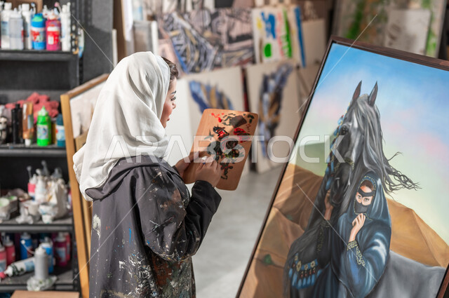 استخدام أدوات و معدات الرسم و التلوين , ورشة عمل فنية , رسامة عربية خليجية سعودية محجبة ترتدي مريلة الرسم و الحجاب تقوم بمزج الألوان المناسبة لإتمام لوحة امرأة عربية مع خيل في الصحراء
