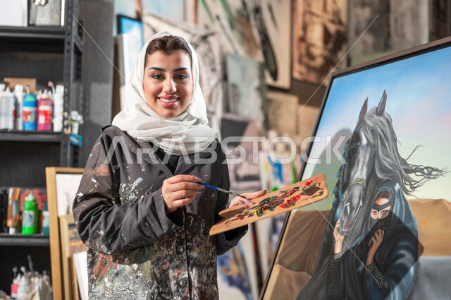 ممارسة هواية الفن التشكيلي في ورشة عمل فنية , رسامة عربية خليجية سعودية مبتسمة ترتدي الثوب مريلة الرسم تمسك بيدها الريشة و  تقف أمام لوحة امرأة عربية مع خيل في الصحراء , النظر إلى الكاميرا بإيماءات الفرح و السرور , دمج الألوان المناسبة لإتمام الرسوم , استخدام أدوات و معدات التلوين