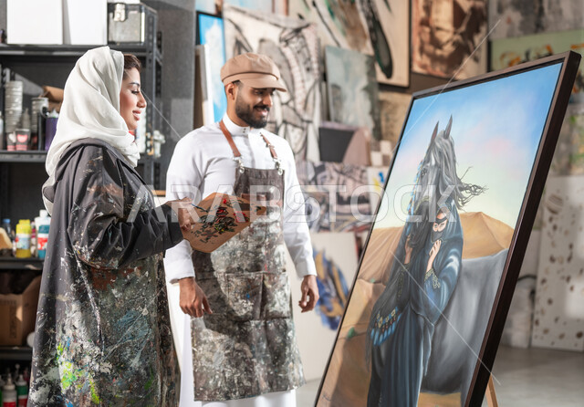 التعاون على تصميم اللوحات المميزة , مفهوم الموهبة و الإبداع , دمج الألوان المناسبة لإتمام الرسوم , رسامان عربيان خليجيان سعوديان يقفان أمام لوحة امرأة عربية مع خيل في الصحراء , ورشة عمل فنية , معرض الرسومات و اللوحات التشكيلية
