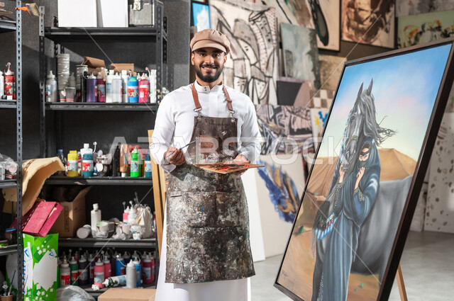 النظر إلى الكاميرا بإيماءات الفرح و السرور , الوقوف أمام لوحة امرأة عربية مع خيل في الصحراء , رسام عربي خليجي سعودي يرتدي الثوب التقليدي و مريلة الرسم , معرض الرسومات و اللوحات التشكيلية في السعودية , ورشة عمل فنية