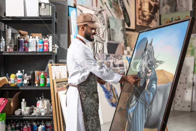 إتمام تفاصيل رسمة امرأة عربية مع خيل في الصحراء , مفهوم الموهبة و الإبداع , رسام عربي خليجي سعودي يرتدي الثوب التقليدي و مريلة الرسم , معرض الرسومات و اللوحات التشكيلية في السعودية , ورشة عمل فنية