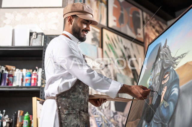 استخدام أدوات الرسم و التلوين , ممارسة الهوايات المفضلة و الفنون اليدوية , رسام عربي خليجي سعودي يرتدي الثوب التقليدي و مريلة الرسم يقوم بإتمام تفاصيل رسمة امرأة عربية مع خيل في الصحراء , معرض الرسومات و اللوحات الفنية التشكيلية في السعودية