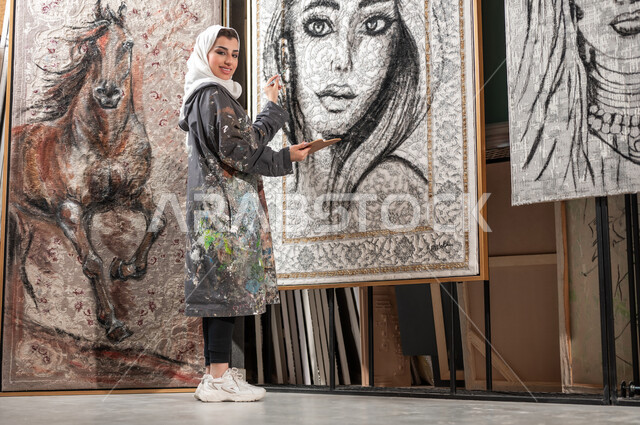 إنجاز المشاريع اللوحات الجدارية المعلقة , معرض الرسوم التشكيلية في السعودية , رسامة عربية خليجية سعودية محجبة ترتدي مريلة الرسم تحمل أدوات و معدات الرسم و التلوين , النظر إلى الكاميرا بإيماءات الفرح و السرور , ورشة عمل فنية