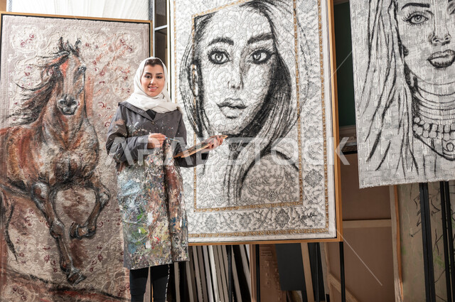 الوقوف أمام اللوحات الجدارية المعلقة , معرض الرسوم التشكيلية في السعودية , رسامة عربية خليجية سعودية محجبة ترتدي مريلة الرسم تحمل أدوات و معدات الرسم و التلوين , النظر إلى الكاميرا بإيماءات الفرح و السرور , ورشة عمل فنية