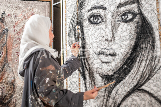 لوحات فنية جدارية معلقة , الشغف بممارسة الفنون اليدوية و الهوايات المفضلة , صورة مقربة من الجانب لرسامة عربية خليجية سعودية محجبة ترتدي مريلة الرسم تقوم بإتمام تفاصيل وجه الامرأة , معرض الرسومات التشكيلية في السعودية